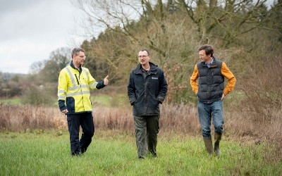 Three men in field, one wearing high vis and walking near the river
